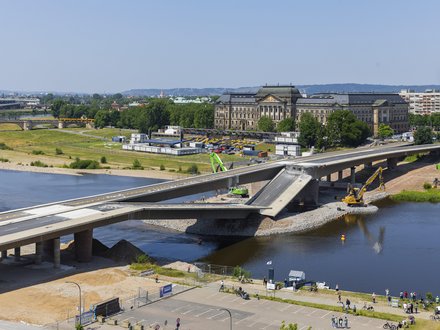 Große Straßenbrücke im Bauzustand über einen Fluss; ein Brückensegment ist abgesenkt, Baumaschinen arbeiten im Wasser und am Ufer, im Hintergrund sind historische Gebäude, Grünflächen und eine Stadtansicht zu sehen.