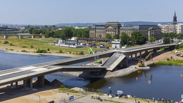 Große Straßenbrücke im Bauzustand über einen Fluss; ein Brückensegment ist abgesenkt, Baumaschinen arbeiten im Wasser und am Ufer, im Hintergrund sind historische Gebäude, Grünflächen und eine Stadtansicht zu sehen.