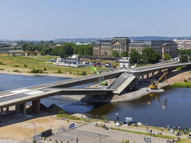 Große Straßenbrücke im Bauzustand über einen Fluss; ein Brückensegment ist abgesenkt, Baumaschinen arbeiten im Wasser und am Ufer, im Hintergrund sind historische Gebäude, Grünflächen und eine Stadtansicht zu sehen.