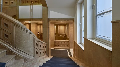 Historisches Treppenhaus mit breiter Steintreppe, Holzgeländer und großen Fenstern mit weißem Rahmen