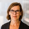 Farbfoto: Nahaufnahme einer Person mit braunen, schulterlangen Haaren und dunkler Brille; neutraler Blick; heller, unscharfer Bürohintergrund.