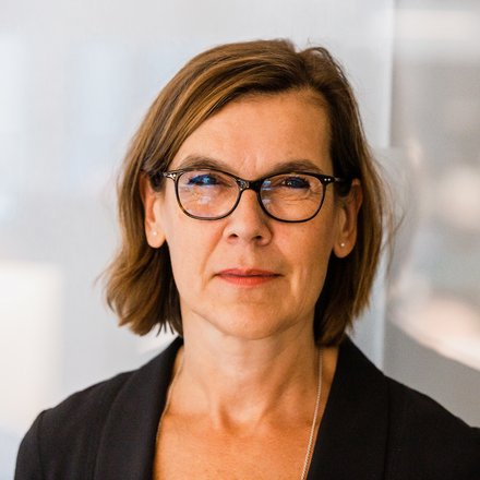 Farbfoto: Nahaufnahme einer Person mit braunen, schulterlangen Haaren und dunkler Brille; neutraler Blick; heller, unscharfer Bürohintergrund.