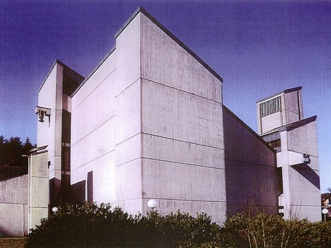 Modernes Betongebäude mit weißer Fassade und gestaffelten kubischen Formen unter blauem Himmel, umgeben von niedrigen Hecken