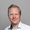 „Porträt eines lächelnden Mannes mit kurzen blonden Haaren und weißem Hemd vor einem grauen Hintergrund.“