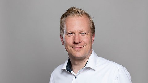 „Porträt eines lächelnden Mannes mit kurzen blonden Haaren und weißem Hemd vor einem grauen Hintergrund.“