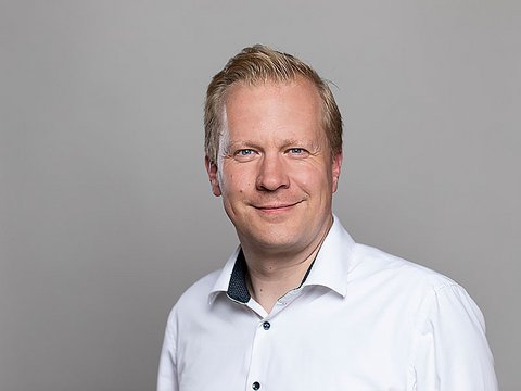 „Porträt eines lächelnden Mannes mit kurzen blonden Haaren und weißem Hemd vor einem grauen Hintergrund.“