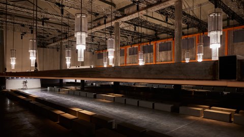 "Großer, leerer Veranstaltungsraum mit langem Laufsteg und vielen gleichmäßig angeordneten, zylindrischen Hängelampen. Betonpfeiler und sichtbare Deckenkonstruktionen verleihen dem Raum einen industriellen Charakter. Sitzblöcke stehen entlang des Laufstegs."