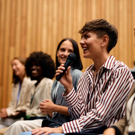 Teilnehmende in einem Vortragssaal sitzen in einer Reihe; im Vordergrund lächelt eine Person im gestreiften Hemd und spricht mit einem Handmikrofon, daneben hören weitere zu.