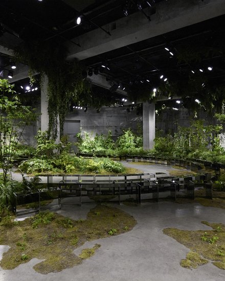 "Ausstellungsraum mit industrieller Architektur, der als Waldlandschaft gestaltet ist. Eine großzügige Halle mit sichtbaren Betondecken und Stahlträgern ist mit üppiger grüner Vegetation gefüllt. Verschiedene Bäume und Sträucher unterschiedlicher Höhen wachsen aus dem Betonboden. Ein gewundener, mit Moos bewachsener Pfad aus Beton führt durch die Installation. Mehrere schwarze Bank-Installationen folgen dem Weg und laden zum Verweilen ein. Die Decke ist mit LED-Spotlights ausgeleuchtet, die das grüne Laub von oben beleuchten. Die Installation verschmilzt die urbane Industriearchitektur mit natürlicher, waldähnlicher Vegetation."
