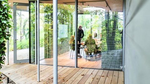 Besprechungsraum in einem gläsernen Pavillon; geöffnete Türen zum Holzdeck, mehrere Personen am Tisch, Spiegelungen der Bäume.