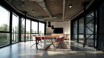 Heller Besprechungsraum mit bodentiefen Fenstern und Sichtbetondecke mit offenen Lüftungsrohren; langer Holztisch, rosafarbene Drehstühle, drei schwarze Pendelleuchten und ein Wandbildschirm im Hintergrund.
