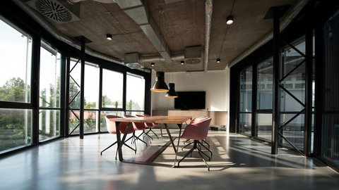 Heller Besprechungsraum mit bodentiefen Fenstern und Sichtbetondecke mit offenen Lüftungsrohren; langer Holztisch, rosafarbene Drehstühle, drei schwarze Pendelleuchten und ein Wandbildschirm im Hintergrund.