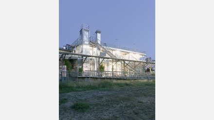 Nachtansicht eines verglasten Anbaus mit außenliegenden Treppen; Innenräume hell erleuchtet, Wiese im Vordergrund.