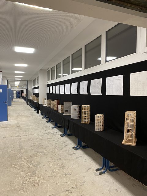 „Blick entlang eines Flurs mit einer langen Ausstellungsreihe von filigranen Holzmodellen und Zeichnungen auf schwarzem Hintergrund.“