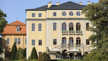 Frontalaufnahme eines großen, sonnig gelben Herrenhauses mit mehreren Stockwerken, hohen Rundbogenfenstern und steinerner Balustrade; im Erdgeschoss Terrasse mit Tischen und großen Sonnenschirmen, davor gepflegte Sträucher und Bäume, klarer blauer Himmel.
