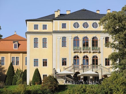 Frontalaufnahme eines großen, sonnig gelben Herrenhauses mit mehreren Stockwerken, hohen Rundbogenfenstern und steinerner Balustrade; im Erdgeschoss Terrasse mit Tischen und großen Sonnenschirmen, davor gepflegte Sträucher und Bäume, klarer blauer Himmel.