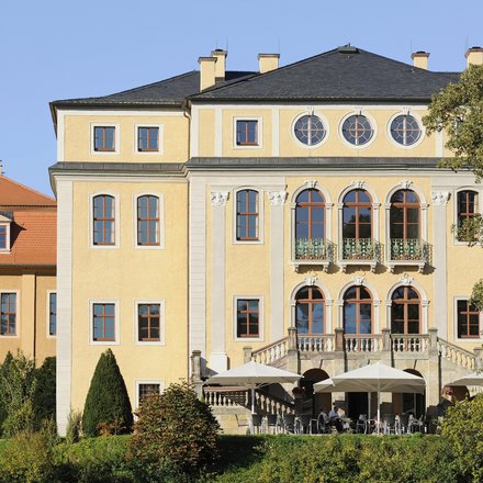 Frontalaufnahme eines großen, sonnig gelben Herrenhauses mit mehreren Stockwerken, hohen Rundbogenfenstern und steinerner Balustrade; im Erdgeschoss Terrasse mit Tischen und großen Sonnenschirmen, davor gepflegte Sträucher und Bäume, klarer blauer Himmel.