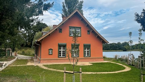 "Orangerotes Einfamilienhaus mit Satteldach und grünen Holzfenstern, umgeben von grüner Wiese und Bäumen. Im Hintergrund ist ein ruhiger See zu sehen. Ein junger Baum mit Schutzvorrichtung steht auf dem Rasen. Links im Bild ein Gartentor mit Backsteinmauer."