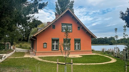 "Orangerotes Einfamilienhaus mit Satteldach und grünen Holzfenstern, umgeben von grüner Wiese und Bäumen. Im Hintergrund ist ein ruhiger See zu sehen. Ein junger Baum mit Schutzvorrichtung steht auf dem Rasen. Links im Bild ein Gartentor mit Backsteinmauer."
