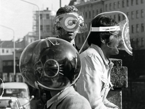 Schwarz-Weiß-Fotografe aus den 1960er Jahren: Zwei Männer tragen futuristische Schutzvorrichtungen mit großen, runden, spiralförmigen Brillengläsern und Kopfbedeckungen. Sie stehen vor einer städtischen Kulisse mit Hochhäusern im Hintergrund. Der Mann links hält eine große metallische Kugel mit spiralförmigen Mustern, der Mann rechts trägt eine flache, rechteckige Vorrichtung. Beide sind in Alltagskleidung gekleidet. Das Bild dokumentiert experimentelle oder künstlerische Visionen von zukünftiger Technologie und Design.