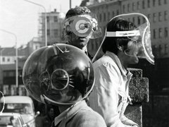 Schwarz-Weiß-Fotografe aus den 1960er Jahren: Zwei Männer tragen futuristische Schutzvorrichtungen mit großen, runden, spiralförmigen Brillengläsern und Kopfbedeckungen. Sie stehen vor einer städtischen Kulisse mit Hochhäusern im Hintergrund. Der Mann links hält eine große metallische Kugel mit spiralförmigen Mustern, der Mann rechts trägt eine flache, rechteckige Vorrichtung. Beide sind in Alltagskleidung gekleidet. Das Bild dokumentiert experimentelle oder künstlerische Visionen von zukünftiger Technologie und Design.