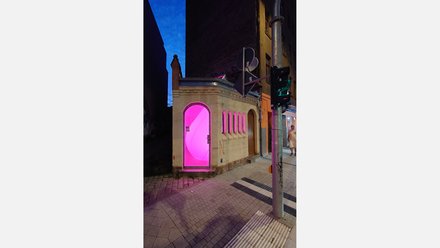 Kleines Eckgebäude „das Bedürfnis“ bei Nacht; der Innenraum leuchtet pink durch den Rundbogen-Eingang und die schmalen Fenster.