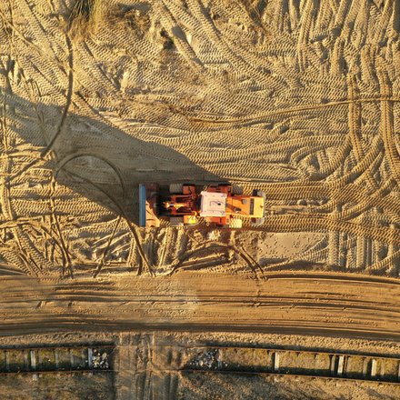 Draufsicht auf eine orangefarbene Baumaschine, die mittig auf weitflächigem Sandboden steht; der Sand ist von zahlreichen Reifen- und Kettenmustern durchzogen, die Maschine wirft einen langen Schatten nach links, am unteren Bildrand ein schmaler Streifen mit Bohlen- oder Verbauelementen.