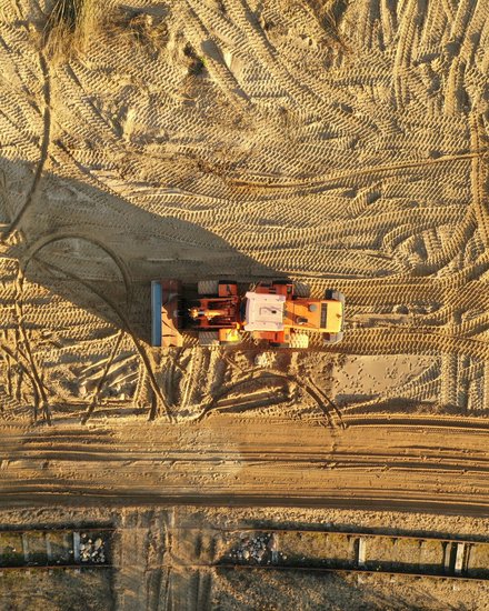 Draufsicht auf eine orangefarbene Baumaschine, die mittig auf weitflächigem Sandboden steht; der Sand ist von zahlreichen Reifen- und Kettenmustern durchzogen, die Maschine wirft einen langen Schatten nach links, am unteren Bildrand ein schmaler Streifen mit Bohlen- oder Verbauelementen.