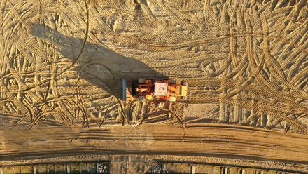 Draufsicht auf eine orangefarbene Baumaschine, die mittig auf weitflächigem Sandboden steht; der Sand ist von zahlreichen Reifen- und Kettenmustern durchzogen, die Maschine wirft einen langen Schatten nach links, am unteren Bildrand ein schmaler Streifen mit Bohlen- oder Verbauelementen.