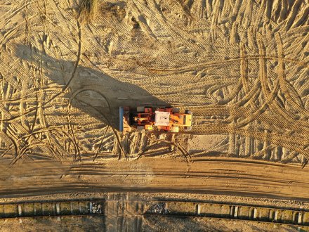 Draufsicht auf eine orangefarbene Baumaschine, die mittig auf weitflächigem Sandboden steht; der Sand ist von zahlreichen Reifen- und Kettenmustern durchzogen, die Maschine wirft einen langen Schatten nach links, am unteren Bildrand ein schmaler Streifen mit Bohlen- oder Verbauelementen.
