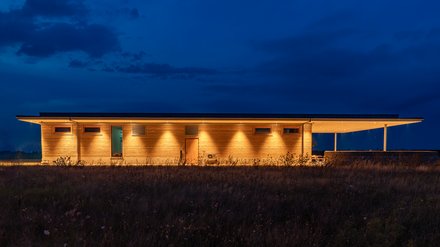 Lange Seitenansicht des Stampflehm-Gebäudes, nachts warm angestrahlt vor tiefblauem Himmel.