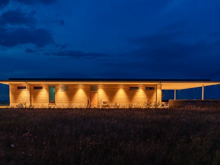 Lange Seitenansicht des Stampflehm-Gebäudes, nachts warm angestrahlt vor tiefblauem Himmel.