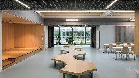 Moderner heller Raum mit organisch geformten Holzbänken in der Mitte, links eine kleine Bühne mit Holzverkleidung, rechts Tische und Stühle, große Fensterfront im Hintergrund