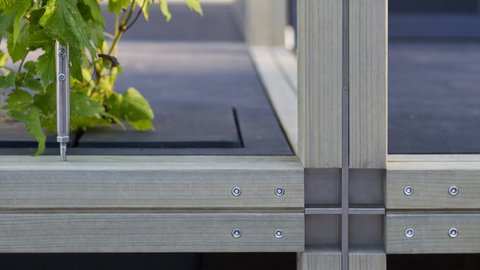 Detailaufnahme eines modularen Holzrahmens mit verschraubten Metallverbindern und Kreuzfuge; links wächst eine grüne Kletterpflanze aus einer eingelassenen Öffnung, geführt an einem gespannten Draht.