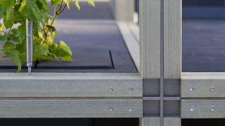 Detailaufnahme eines modularen Holzrahmens mit verschraubten Metallverbindern und Kreuzfuge; links wächst eine grüne Kletterpflanze aus einer eingelassenen Öffnung, geführt an einem gespannten Draht.