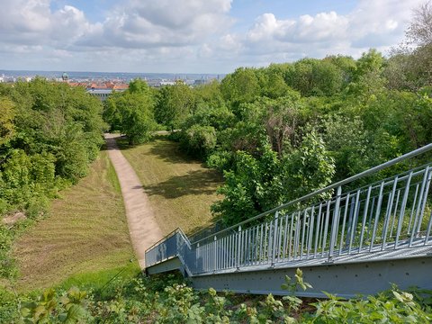 „Blick von einer Metalltreppe auf einen grünen Parkhang. Ein schmaler, geschwungener Fußweg führt zwischen Bäumen und Sträuchern hinab in Richtung Stadt. Im Hintergrund sind Dächer und Kirchtürme unter einem wolkigen blauen Himmel zu sehen.“