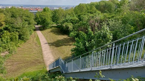„Blick von einer Metalltreppe auf einen grünen Parkhang. Ein schmaler, geschwungener Fußweg führt zwischen Bäumen und Sträuchern hinab in Richtung Stadt. Im Hintergrund sind Dächer und Kirchtürme unter einem wolkigen blauen Himmel zu sehen.“