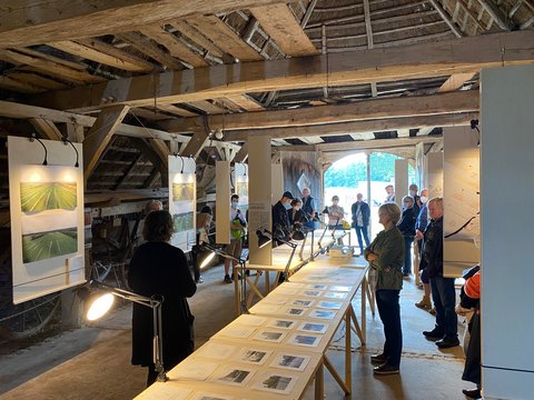 Eine Gruppe besucht eine Ausstellung in einer historischen Scheune. Die Teilnehmenden stehen zwischen beleuchteten Tafeln und langen Tischen mit Fotografien, Karten und Texten.