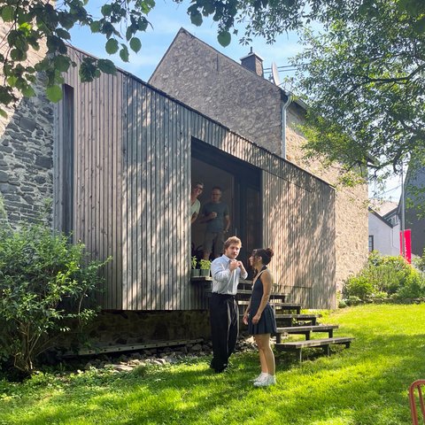 „Zwei Personen unterhalten sich auf den Stufen eines modernen Holzanbaus, der an ein älteres Steinhaus anschließt, umgeben von einem sonnigen, grünen Garten.“
