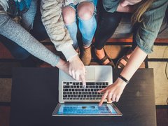 Drei Personen, die gemeinsam auf ein Laptop und ein Tablet zeigen, während sie in einer entspannten Umgebung auf einem Tisch sitzen.