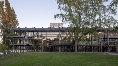 Modernes, mehrstöckiges Bürogebäude mit großer Glasfassade und umlaufendem Balkon vor Rasenfläche und Bäumen