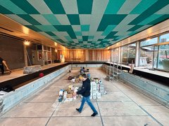 „Baustelle eines Hallenbads: Ein Mann geht durch das leere Betonbecken, über dem sich eine markante Decke mit einem geometrischen Muster in Weiß-, Grün- und Türkistönen erstreckt.“