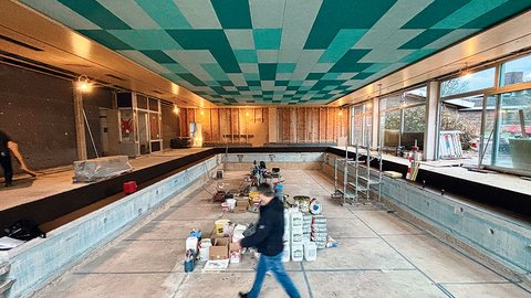 „Baustelle eines Hallenbads: Ein Mann geht durch das leere Betonbecken, über dem sich eine markante Decke mit einem geometrischen Muster in Weiß-, Grün- und Türkistönen erstreckt.“
