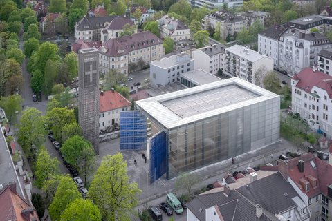 Luftaufnahme eines modernen Gebäudes mit weißer Metallgitterfassade und Flachdach in urbaner Wohnumgebung, daneben ein Gebäude mit blauer Glasfassade, umgeben von klassischen Wohnhäusern mit roten Ziegeldächern, grünen Bäumen und gepflasterten Plätzen
