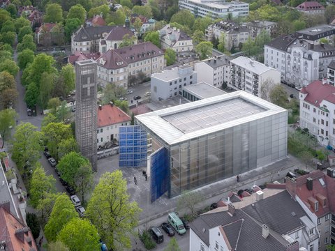 Luftaufnahme eines modernen Gebäudes mit weißer Metallgitterfassade und Flachdach in urbaner Wohnumgebung, daneben ein Gebäude mit blauer Glasfassade, umgeben von klassischen Wohnhäusern mit roten Ziegeldächern, grünen Bäumen und gepflasterten Plätzen