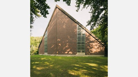 Außenansicht einer modernen Backsteinkirche mit markantem Satteldach, hohen Fensterbändern und parkähnlicher Grünfläche.