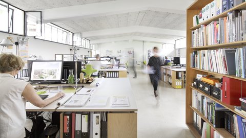Heller Büroraum mit Arbeitsplätzen, Bücherregalen und großen Fenstern.