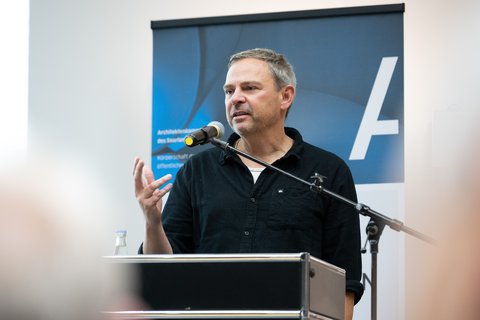 Mann mittleren Alters steht hinter einem Rednerpult und spricht ins Mikrofon; er trägt ein dunkles Hemd, gestikuliert mit der rechten Hand und blickt leicht nach rechts; hinter ihm ein blaues Roll‑up mit weißem Schriftzeichen als Hintergrund.