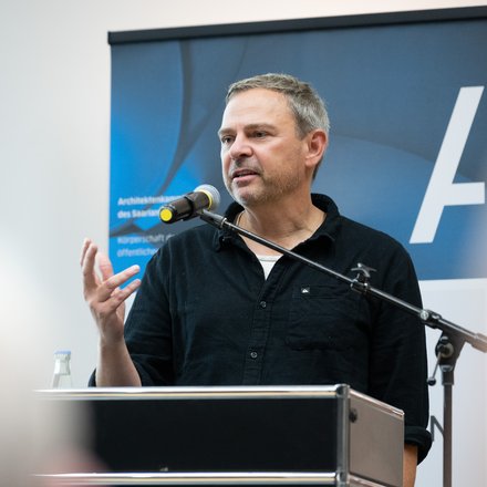 Mann mittleren Alters steht hinter einem Rednerpult und spricht ins Mikrofon; er trägt ein dunkles Hemd, gestikuliert mit der rechten Hand und blickt leicht nach rechts; hinter ihm ein blaues Roll‑up mit weißem Schriftzeichen als Hintergrund.