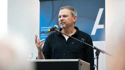 Mann mittleren Alters steht hinter einem Rednerpult und spricht ins Mikrofon; er trägt ein dunkles Hemd, gestikuliert mit der rechten Hand und blickt leicht nach rechts; hinter ihm ein blaues Roll‑up mit weißem Schriftzeichen als Hintergrund.